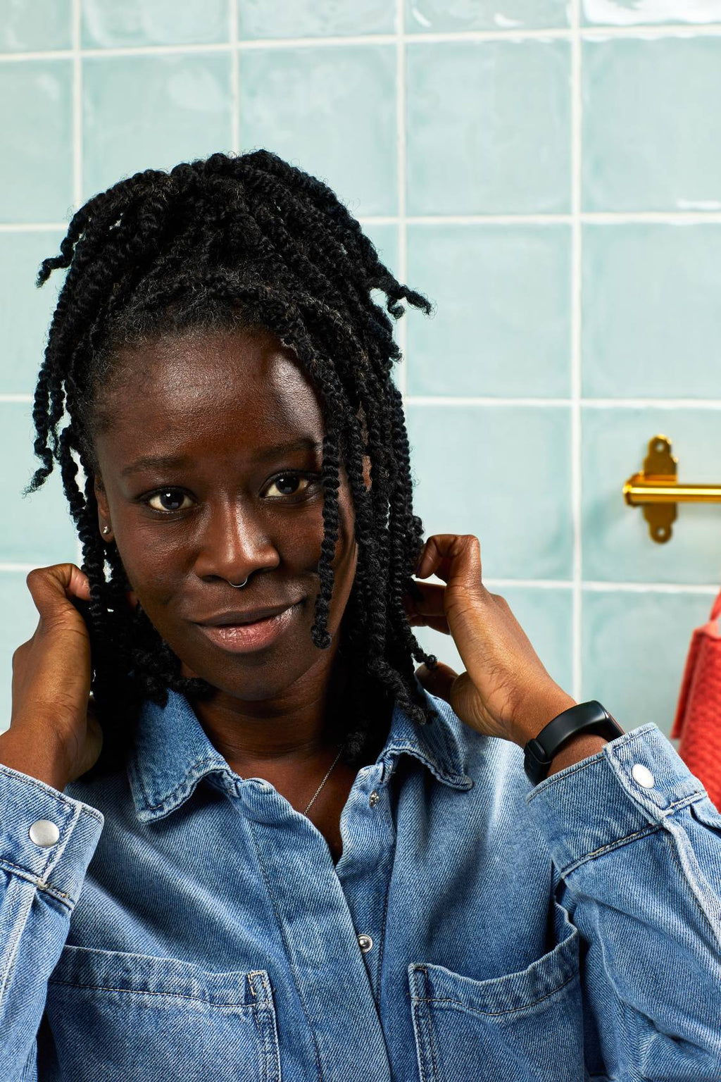 A smiling person with braided hair poses against a backdrop of light blue bathroom tiles. Wearing a denim shirt, they hold the ends of their hair with both hands, looking directly at the camera.
